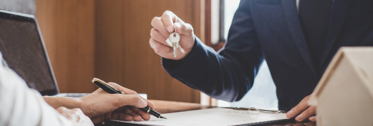 Une personne tend une clé à une autre en train de signer un papier représentant un locataire qui signe son contrat de location avec le gestionnaire locatif